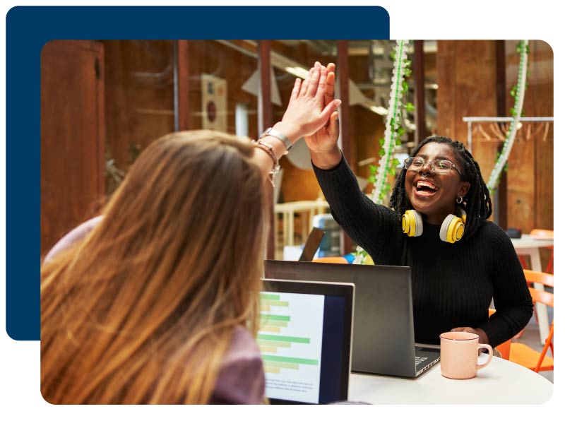 Portrait of two multiracial work colleagues giving each other high-five to express success while sitting at their workstations.