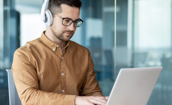 A young man sits at a workplace in the office, works, studies on a laptop with headphones. Listens to the webinar, music, course.