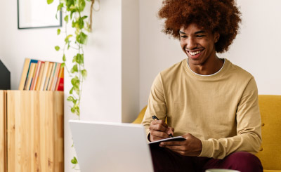 Young adult african american man writing on notebook while watching online training class on laptop computer. E-learning and education online concept