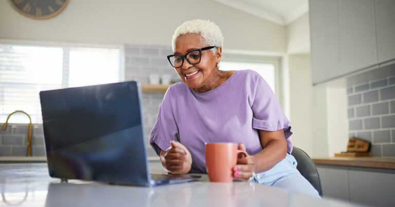 An elderly woman with short white hair and glasses smiles while looking at her laptop in a bright kitchen. She holds a peach-colored mug.