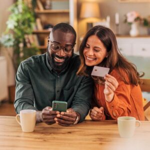Smiling couple enjoying time together at their dining table, using a smartphone and credit card for online shopping and making secure payments in the comfort of their home