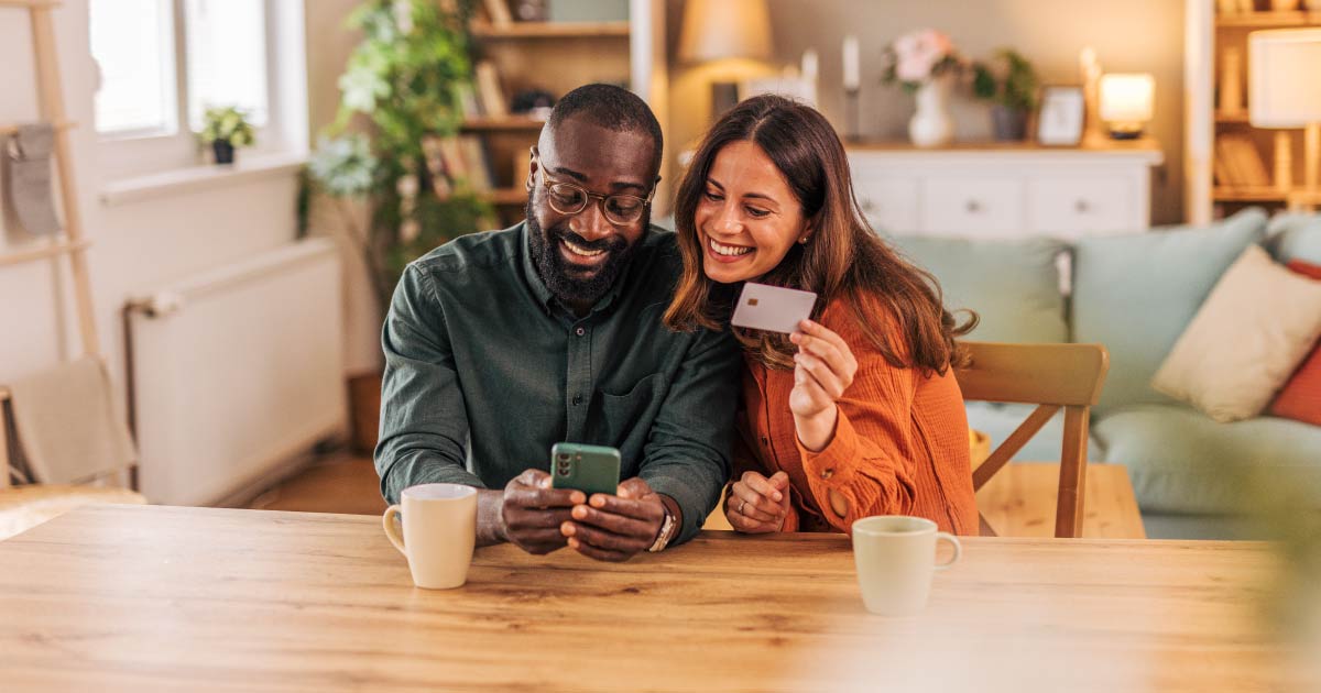 Smiling couple enjoying time together at their dining table, using a smartphone and credit card for online shopping and making secure payments in the comfort of their home