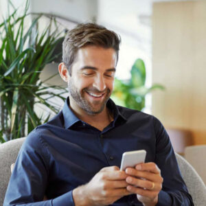 Smiling businessman text messaging on smart phone. Male executive is sitting on couch. He is using mobile phone at office.