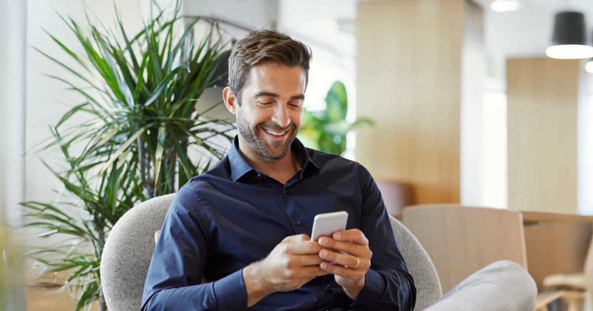 Smiling businessman text messaging on smart phone. Male executive is sitting on couch. He is using mobile phone at office.