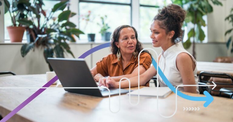 Young adult and mature women, dressed in professional attire, engage in collaborative work at a laptop in a contemporary office