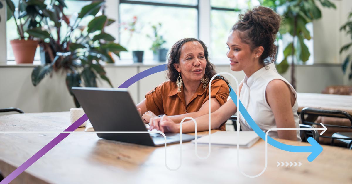 Young adult and mature women, dressed in professional attire, engage in collaborative work at a laptop in a contemporary office