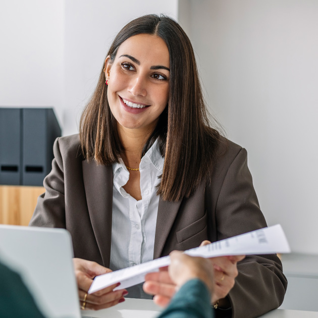 A woman in a brown blazer sits at a desk, smiling and holding a document while discussing grants with another person from a nonprofit