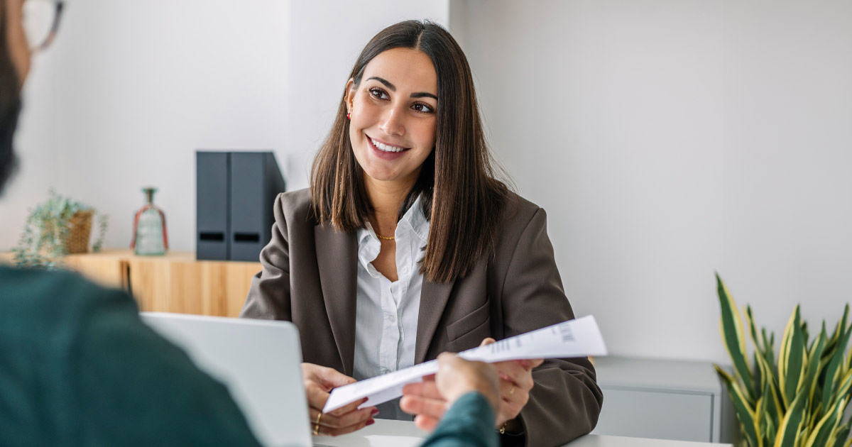 A woman in a brown blazer sits at a desk, smiling and holding a document while discussing grants with another person from a nonprofit