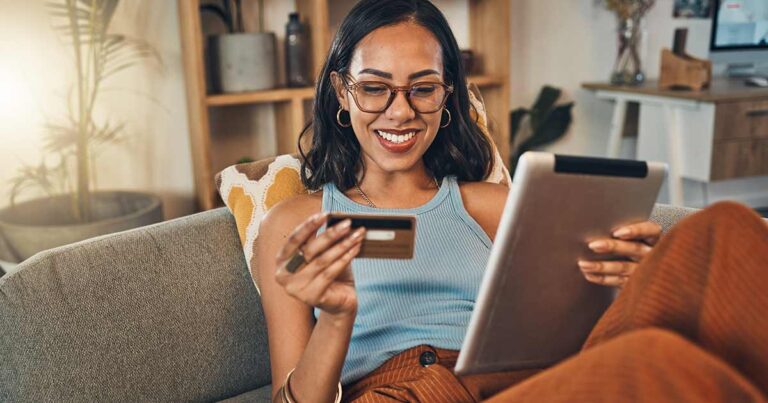 Smiling mixed race woman using credit card for ecommerce on digital tablet at home. Happy hispanic sitting alone on living room sofa, using technology for ebanking. Relaxing, ordering, buying online