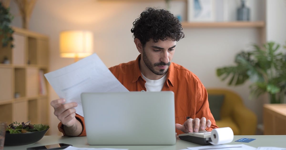 Front view of a young man, dark haired and bearded, dressed in casual clothes, sitting in the living room of his apartment reviewing bills with a calculator and laptop.
