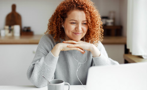 Female freelancer having video conference with her employer discussing new project details, listening to him attentively in headphones smiling at web camera, putting folded hands under chin